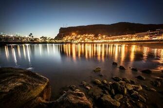 Black Rocks On Body Of Water Near City Lights Under Blue Sky